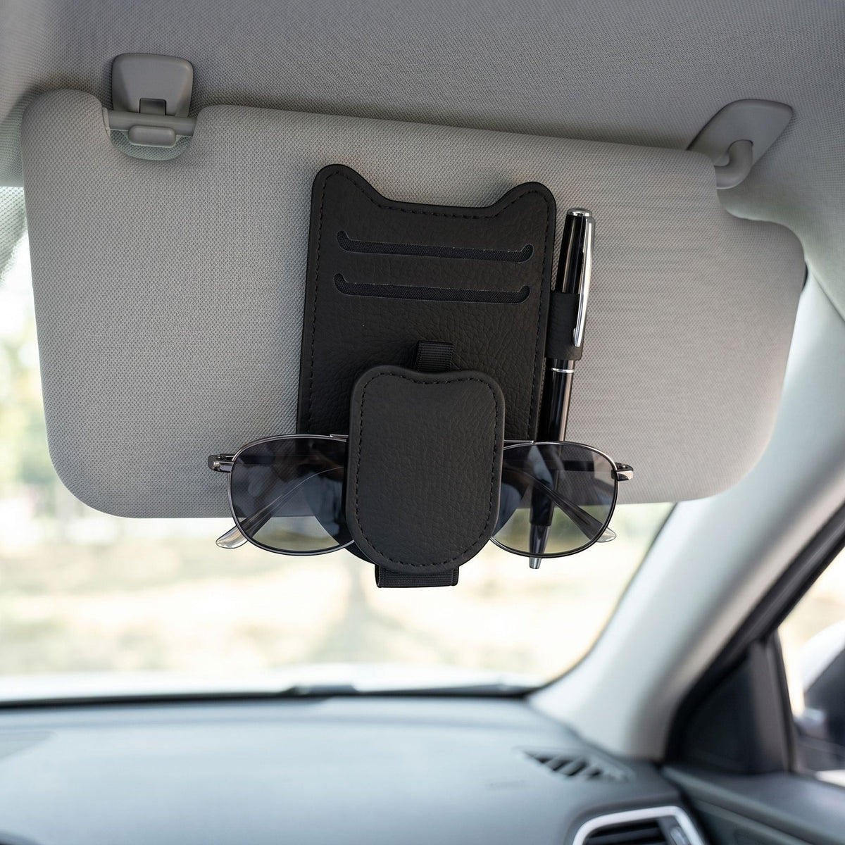 Lifestyle scene showing magnetic sunglasses holder clipped to car sun visor with sunglasses inside, in a bright car interior.