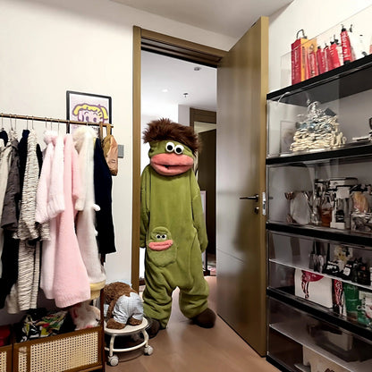 A person wearing a green monster fleece onesie pajama from zero2cart with a brown hair tuft and a pink mouth pocket.